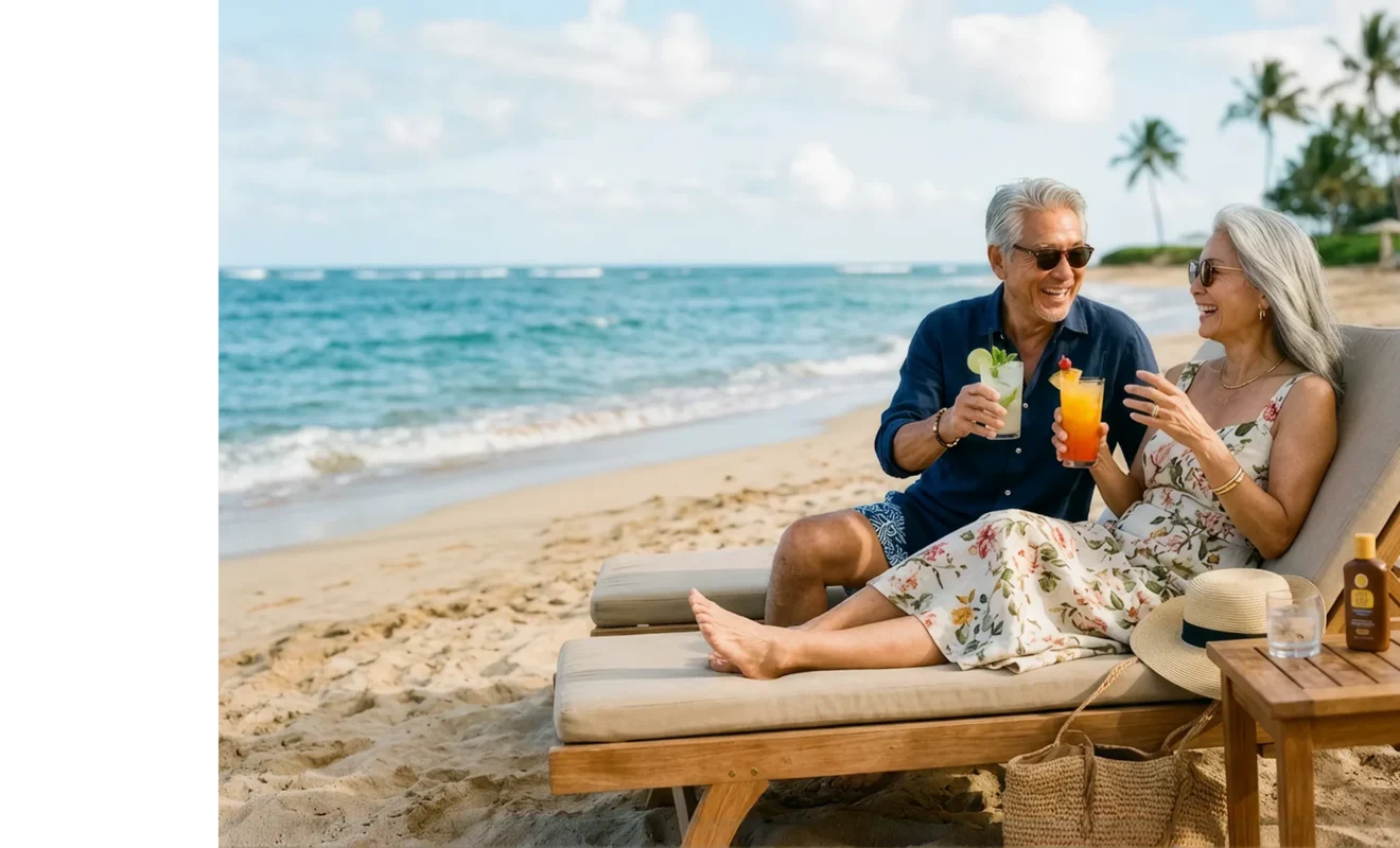 Happy retired couple relaxing on beach loungers with cocktails, laughing together by the ocean.